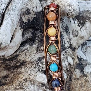 Pode incluir: Um bracelete envolto em fio de cobre com sete pedras preciosas coloridas, representando os sete chakras. O bracelete é exibido em um pedaço de madeira à deriva.