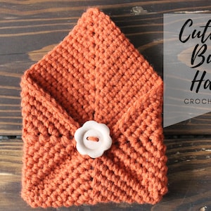 May include: A crocheted button hanger in a burnt orange colour with a white button in the centre. The hanger is folded into a triangle shape and is sitting on a wooden surface. The text "Cute as a Button Hanger CROCHET PATTERN" is visible in the image.