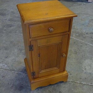 May include: A light brown wooden bedside cabinet featuring a rectangular top, one upper drawer with a single round knob, and a lower cupboard door with a matching round knob and visible metal hinges, supported by a scalloped base design.