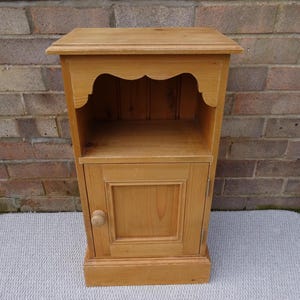 May include: A light-coloured wooden bedside cabinet with a single door and a small shelf. The cabinet has a scalloped design above the shelf and a round wooden knob on the door. The cabinet is approximately 76 cm tall.