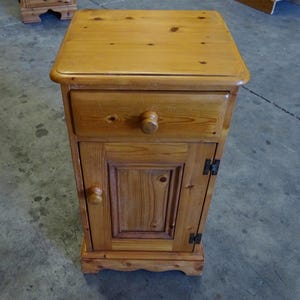 May include: A light brown wooden bedside cabinet featuring a rectangular top, one drawer with a single round wooden knob, and a lower cupboard door with a matching round knob, visible wood grain, and dark metal hinges on the right side of the door.