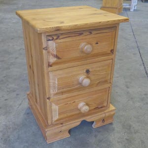 May include: A light brown wooden bedside cabinet featuring three stacked drawers, each fitted with a single round wooden knob. The piece has a rectangular top surface, visible wood grain patterns, and a base with a decorative cutout design.
