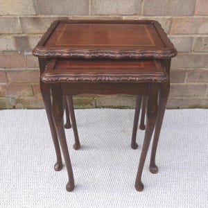 May include: A pair of nesting tables in a dark brown hue. The tables feature square tops with ornate edges and carved patterns. The slender, curved legs add to the design. The tables are made of wood.