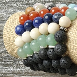 May include: A stack of colorful beaded bracelets with a variety of stones, including red, blue, white, green, and black. The bracelets are made with lava rock beads and are stacked on a burlap background.