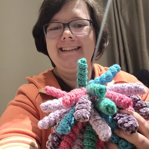 May include: A person is holding a colourful crocheted pom-pom with multiple strands of yarn in shades of purple, pink, teal and green.