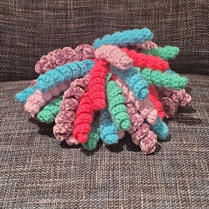 May include: A colourful crocheted yarn bundle in shades of pink, purple, teal and green. The yarn is arranged in a loose, fluffy pile.