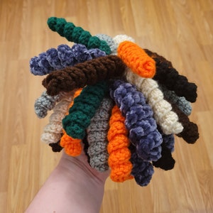 May include: A collection of crocheted yarn strands in various colours, including green, blue, brown, orange, and white. The strands are twisted together to create a unique and textured look.