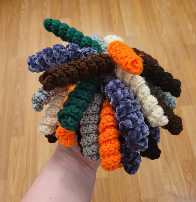 May include: A collection of crocheted yarn strands in various colours, including green, blue, brown, orange, and white. The strands are twisted together to create a unique and textured look.