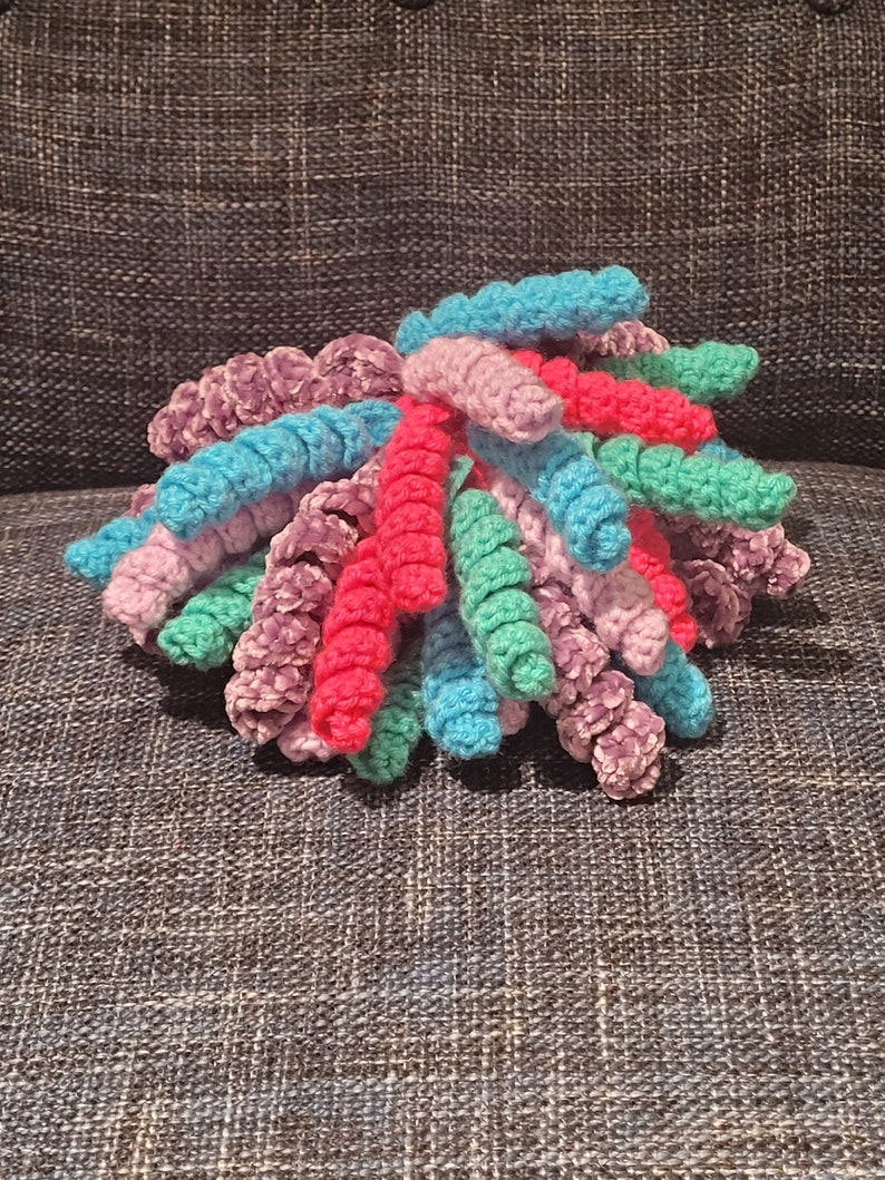 May include: A colourful crocheted yarn bundle with pink, purple, teal and green yarn strands. The yarn is arranged in a loose, fluffy pile.