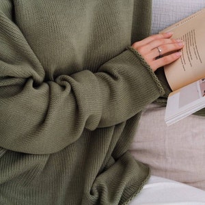 May include: A loose-fitting, olive green knitted sweater with a wide, round neckline. The sweater is made of a soft, comfortable material and has a relaxed fit.