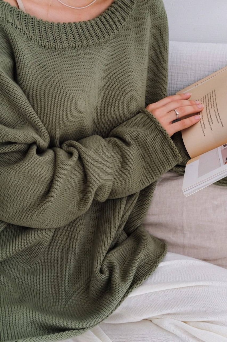 May include: A loose-fitting, olive green knitted sweater with a wide, round neckline. The sweater is made of a soft, comfortable material and has a relaxed fit.