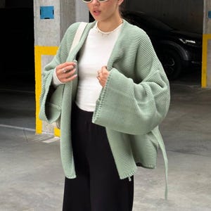 Puede incluir: Un c&aacute;rdigan de punto verde salvia con mangas largas y un cintur&oacute;n de lazo. El c&aacute;rdigan est&aacute; abierto por delante y tiene un corte holgado. La modelo lleva una camiseta blanca y pantalones negros.