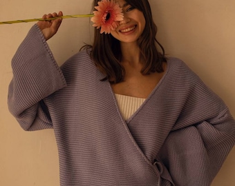 May include: A woman wearing a light purple wrap cardigan with long sleeves and a tie at the waist. She is holding a pink gerbera daisy in front of her face and smiling.