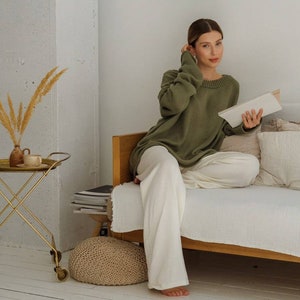 May include: A woman wearing a green sweater and white pants sits on a couch and reads a book. She is in a room with a white wall, a gold side table, and a round mirror hanging on the wall.