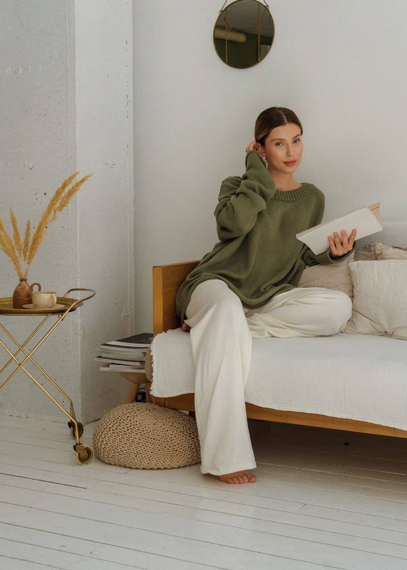 May include: A woman wearing a green sweater and white pants sits on a couch and reads a book. She is in a room with a white wall, a gold side table, and a round mirror hanging on the wall.