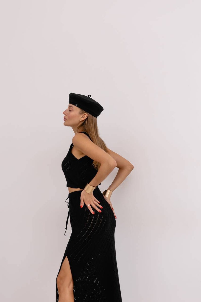 May include: A woman wearing a black beret, a black sleeveless top, and a black skirt with a side slit. She is wearing gold bracelets on both wrists.
