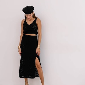May include: A woman wearing a black knit top and skirt set with a black beret. The skirt has a slit on the side and the top has a v-neckline. She is wearing gold bracelets and gold heeled sandals.