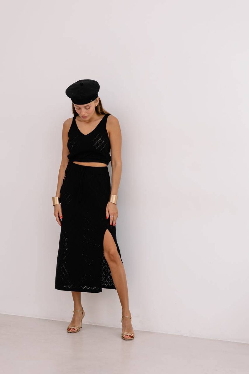 May include: A woman wearing a black knit top and skirt set with a black beret. The skirt has a slit on the side and the top has a v-neckline. She is wearing gold bracelets and gold heeled sandals.