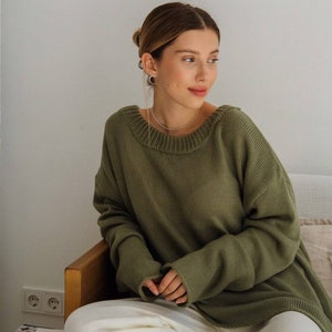 May include: A woman wearing a loose-fitting, olive green sweater with long sleeves and a round neckline. She is sitting on a couch with white pants and has her hair pulled back.