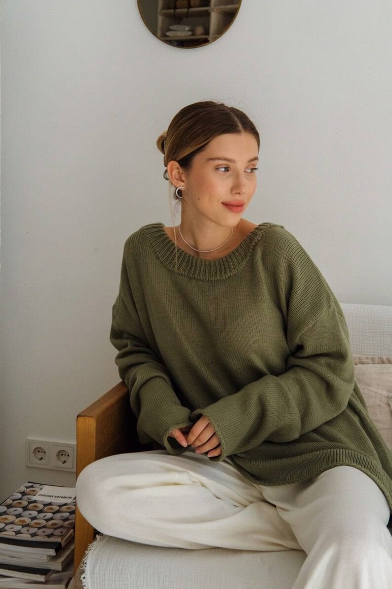 May include: A woman wearing a loose-fitting, olive green sweater with long sleeves and a round neckline. She is sitting on a couch with white pants and has her hair pulled back.