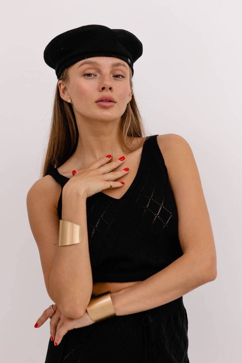 May include: A woman wearing a black beret, a black sleeveless top with a diamond pattern, and a gold bracelet. She is wearing a black skirt with a similar diamond pattern.