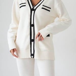 May include: A white cardigan with black trim and buttons. The cardigan has a v-neck and a patch pocket on the left side.