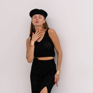 May include: A woman wearing a black beret, a black crop top with a diamond pattern, and a black skirt with a slit. She is wearing gold bracelets on both wrists.