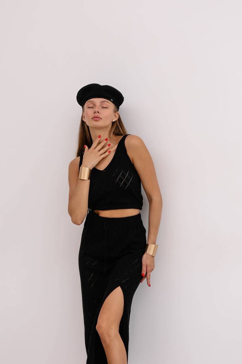 May include: A woman wearing a black beret, a black crop top with a diamond pattern, and a black skirt with a slit. She is wearing gold bracelets on both wrists.