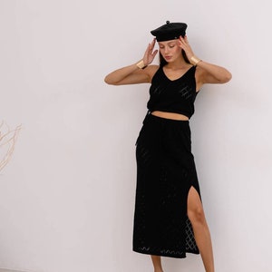May include: A woman wearing a black knit top and skirt with a side slit. She is wearing a black beret and gold bracelets.