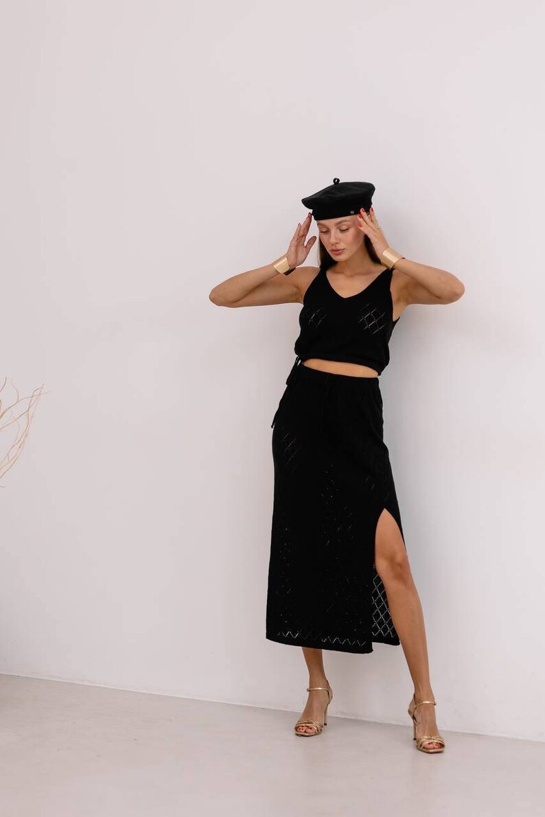 May include: A woman wearing a black knit top and skirt with a side slit. She is wearing a black beret and gold bracelets.
