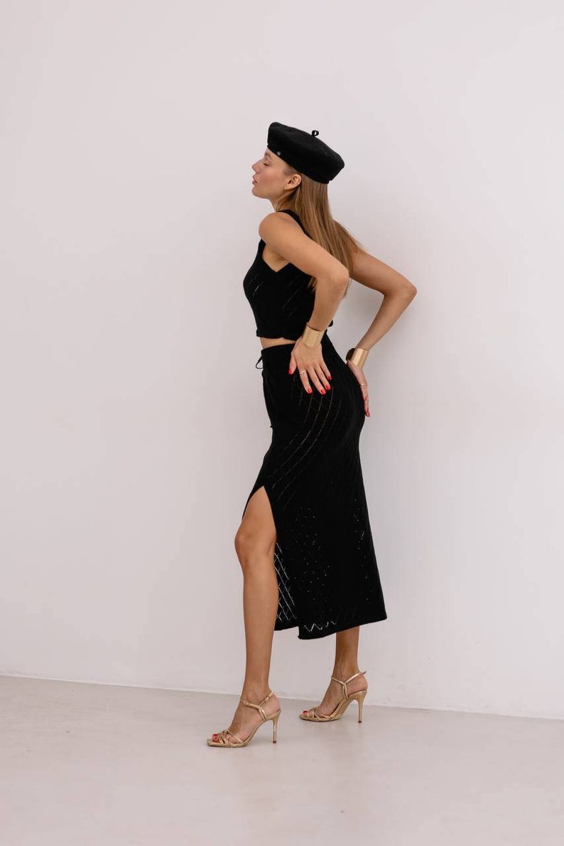 May include: A woman wearing a black knit top and skirt set with a black beret. She is wearing gold bracelets and gold heeled sandals.