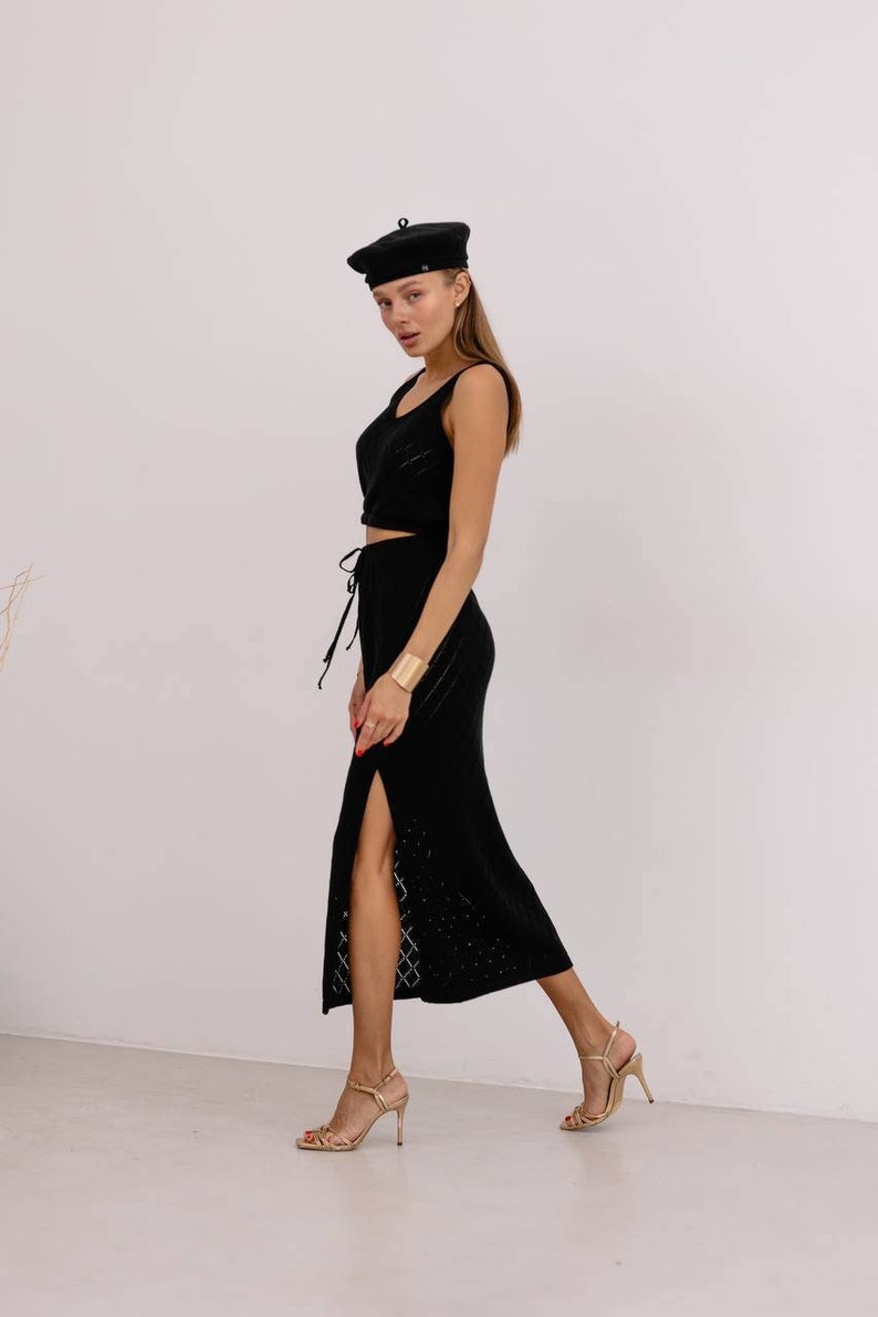 May include: A woman wearing a black knit crop top and matching skirt with a side slit. She is wearing a black beret and gold heels.
