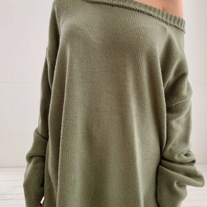 May include: A loose-fitting, oversized green sweater with a slightly off-the-shoulder neckline. The sweater has a relaxed fit and a slightly uneven hemline.