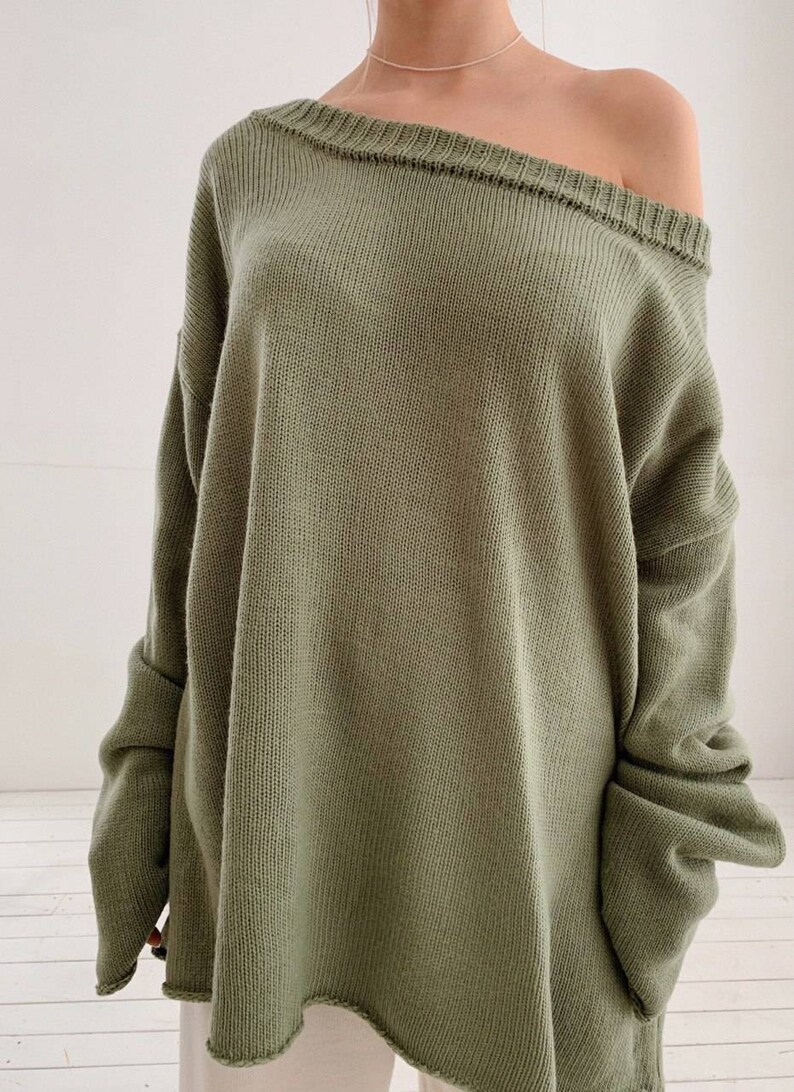 May include: A loose-fitting, oversized green sweater with a slightly off-the-shoulder neckline. The sweater has a relaxed fit and a slightly uneven hemline.