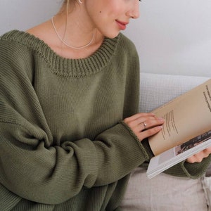 May include: A woman wearing a green knitted sweater with a relaxed fit and a round neckline. She is sitting on a couch and reading a book.