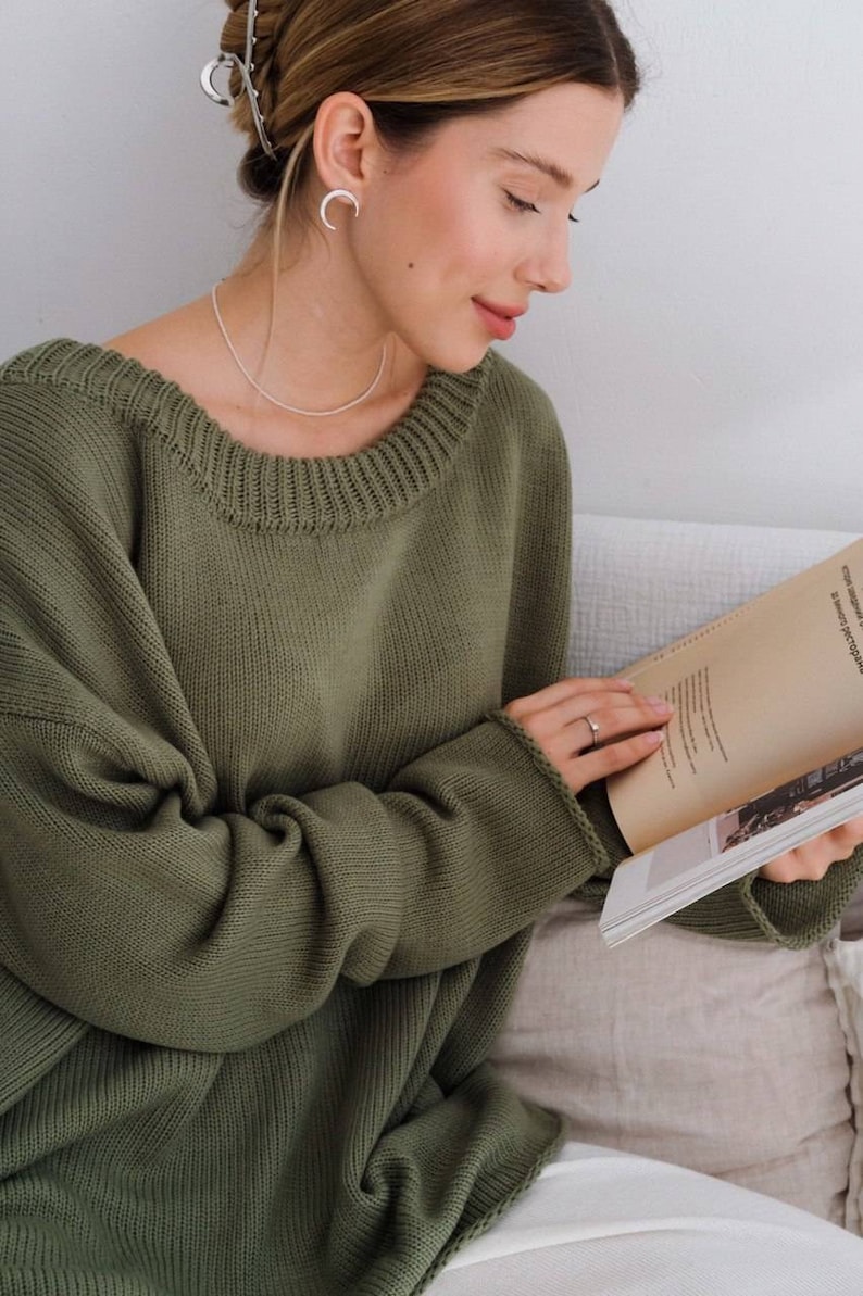 May include: A woman wearing a green knitted sweater with a relaxed fit and a round neckline. She is sitting on a couch and reading a book.