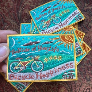 Peut inclure: Un lot de patchs brodés avec un fond turquoise, des fleurs jaunes, un soleil, une lune et un vélo. Le texte sur les patchs est "The Ride of Your Life" et "Bicycle Happiness".