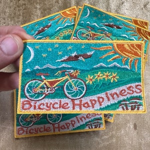 May include: Four embroidered patches with a yellow border, a turquoise background, and the text "Bicycle Happiness". The patches feature a bicycle, a sun, a moon, and a bird.