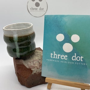 May include: A handmade pottery cup with a green and white glaze, sitting on a brick. A business card on a small wooden easel displays the text "three dot HANDMADE HEIRLOOM POTTERY". A sticker with the same logo is also visible.