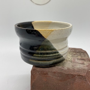 May include: A handmade ceramic bowl with a unique color-block design. The bowl is divided into sections of black, off-white, and tan. The bowl has a textured, ribbed exterior and a smooth interior. The bowl is sitting on a red brick. The logo "three dot" is in the background.