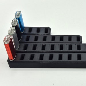 Könnte beinhalten: Schwarzer, mehrstufiger Präsentationsständer mit rechteckigen Schlitzen, der rote, schwarze und blaue Feuerzeuge hält. Der Ständer besteht aus einem mattschwarzen Material und ist in einem absteigenden Treppendesign angeordnet.