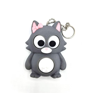 May include: A gray cat-shaped keychain with a pink bow, ears, and nose. The cat has large, wide-eyed black pupils and a white belly. A silver chain and ring are attached to the top. The keychain is made of a smooth, possibly rubbery material.