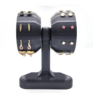 May include: A black earring display stand with two rotating sides. The stand holds various earrings, including gold-colored studs, dangles, and heart-shaped designs. The earrings are displayed against a black background.