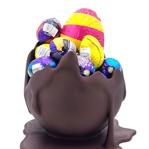 Puede incluir: Un cuenco de chocolate marrón oscuro en forma de huevo lleno de huevos de chocolate envueltos en papel de aluminio. Los colores del papel de aluminio son amarillo, morado, azul y rosa. El cuenco se apoya en una base marrón oscuro.