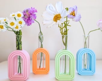 May include: Four colourful bud vases in pink, orange, green, and blue, each holding a single stem of flowers. The vases have an arched design and are made of a solid material. The flowers include white daisies, purple, and white blooms.