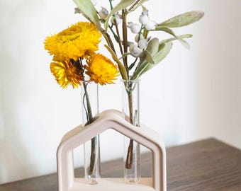 May include: A pale pink house-shaped vase with two clear glass tubes, displaying yellow and white flowers. The vase is on a wooden surface. The flowers provide a splash of colour and natural charm to the decorative item. The vase is approximately 15 cm wide.
