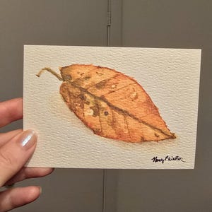 May include: A rectangular watercolor painting on textured paper depicting a single fallen leaf in shades of orange, brown, and yellow. The leaf features visible veins and dark spots. The artist's signature, Nancy P Waller, is written in black ink in the corner.