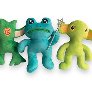 May include: Three plush toys: a green monster with red eyes, a blue frog holding a silver wand, and a green creature with large orange ears.