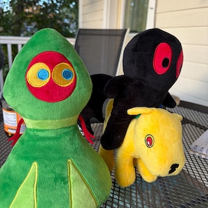 May include: Three plush toys on a metal table. A green plush with a red and yellow face and blue eyes. A black plush with red eyes is riding a yellow dog plush. The toys are soft and colorful.