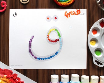 May include: A rainbow-colored Arabic letter 'Qaf' is drawn on a white sheet of paper. The letter is outlined with dots of paint in different colors. There are paint bottles, a paint palette, and cotton swabs in the background.